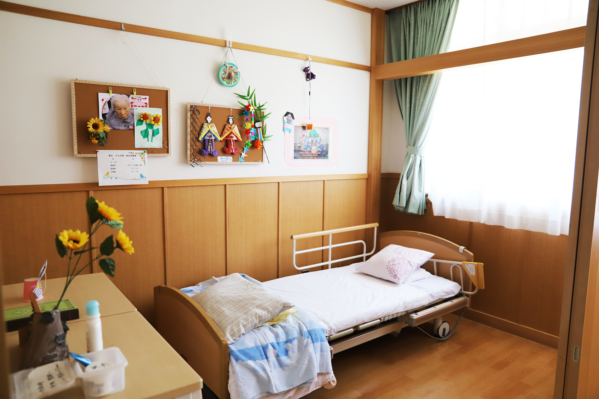 木調の壁と家具で温かみのある居室。ベッドと窓、掲示板で落ち着いた空間。